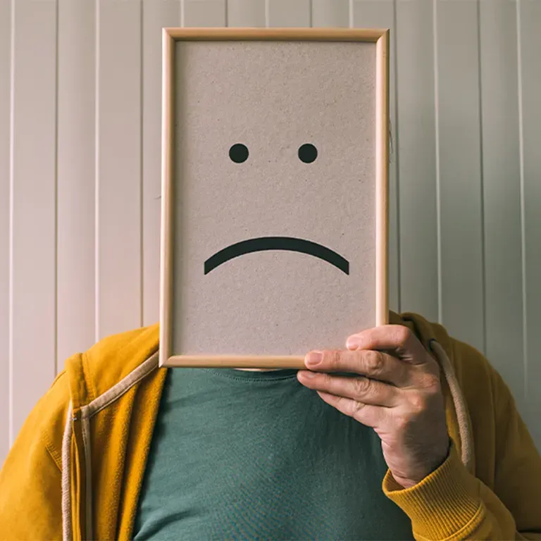 Man holding sign in front of his face, displaying sad face emoticon.